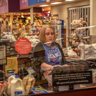 A grocery store clerk rings up items behind a plexiglass barrier
