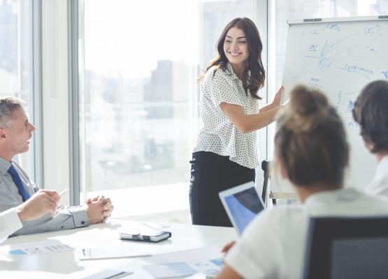 A woman points to a white board while explaining something in an office room